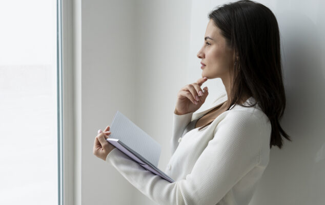 Woman holding a notebook and thinking, representing the process of coming up with self-evaluation examples