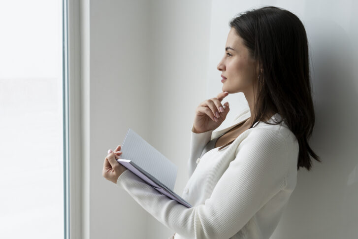 Woman holding a notebook and thinking, representing the process of coming up with self-evaluation examples
